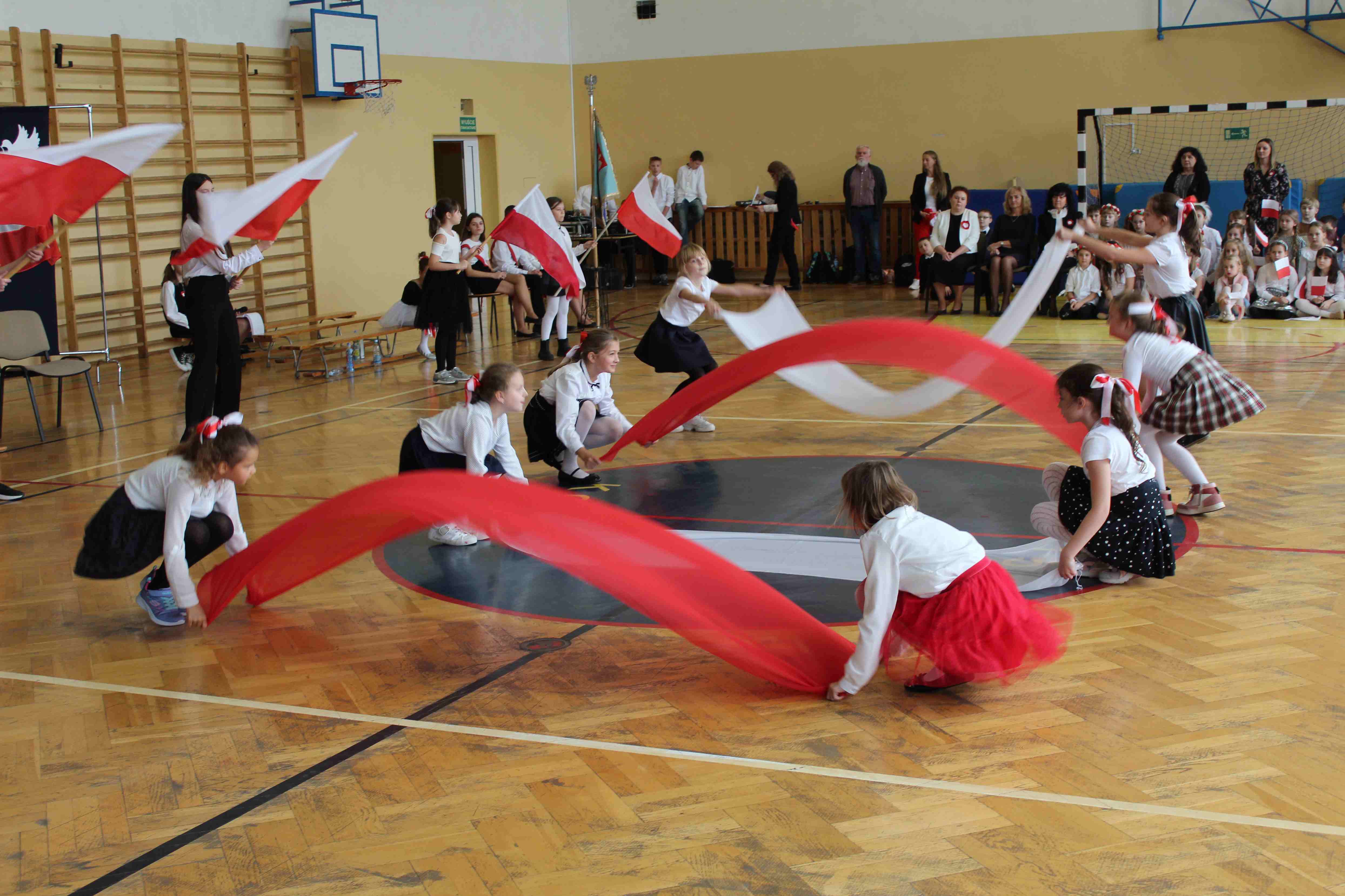 Dzieci tańczą z biało czerwonymi szarfami podczas apelu szkolnego